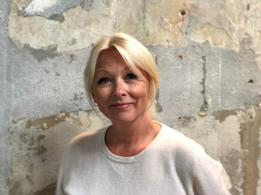 Portrettbilde av Siri Nodland, generalsekretær i Norges Innsamlingsråd. Hun har på seg en lys tynn strikkegenser og står mot en murvegg.