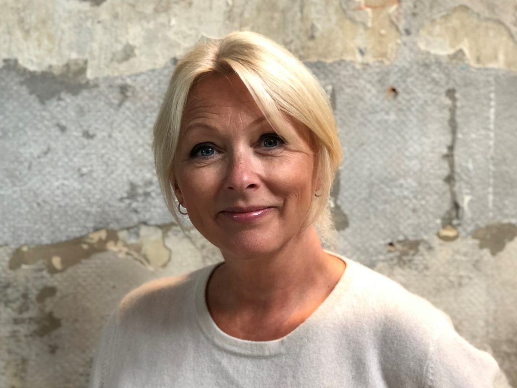 Portrettbilde av Siri Nodland, generalsekretær i Norges Innsamlingsråd. Hun har på seg lys genser og står mot en murvegg.