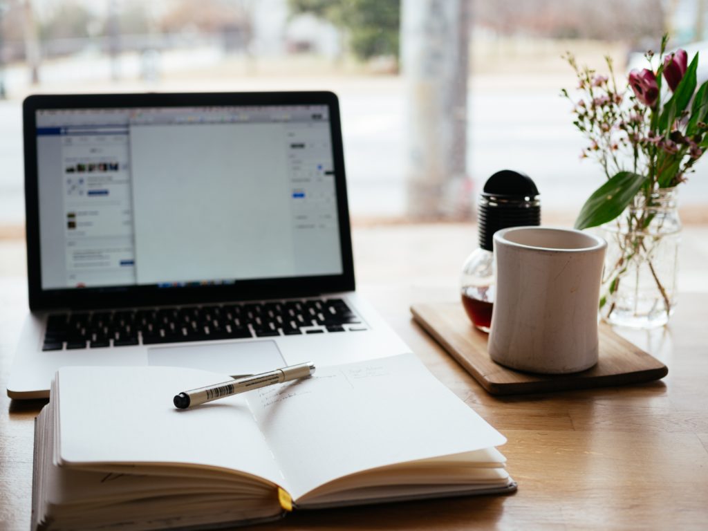 Bilde av et bord med pc, notatblokk og kaffekopp. Skal illustrere at en person sitter og jobber.