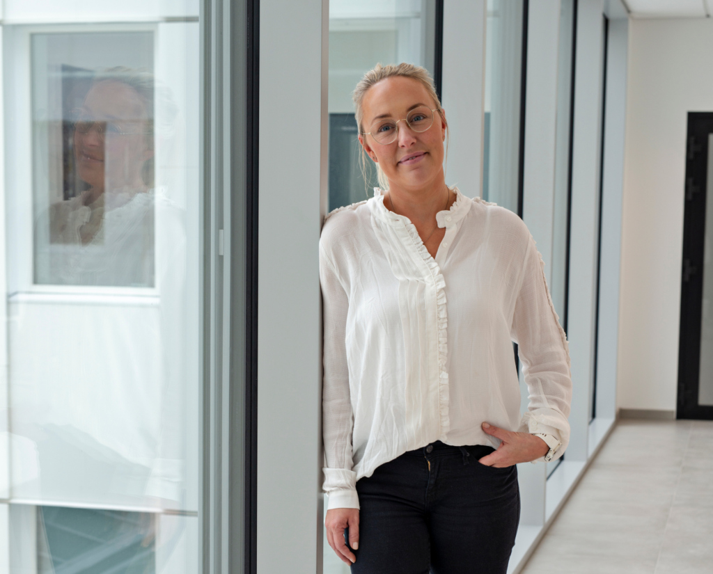Portrettbilde av Marie Sanne Hval. Hun har på seg hvit bluse og sort bukse. Står i kontorlandskap. 