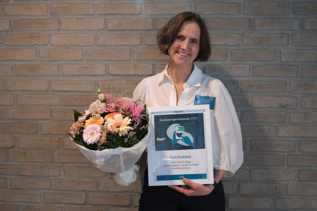 Portrettbilde av Marit Sophie Egge, aksjonsleder i Kreftforeningen. Hun har på seg hvit bluse og mørk bukse. Hun holder en blomsterbukett og et diplom. 